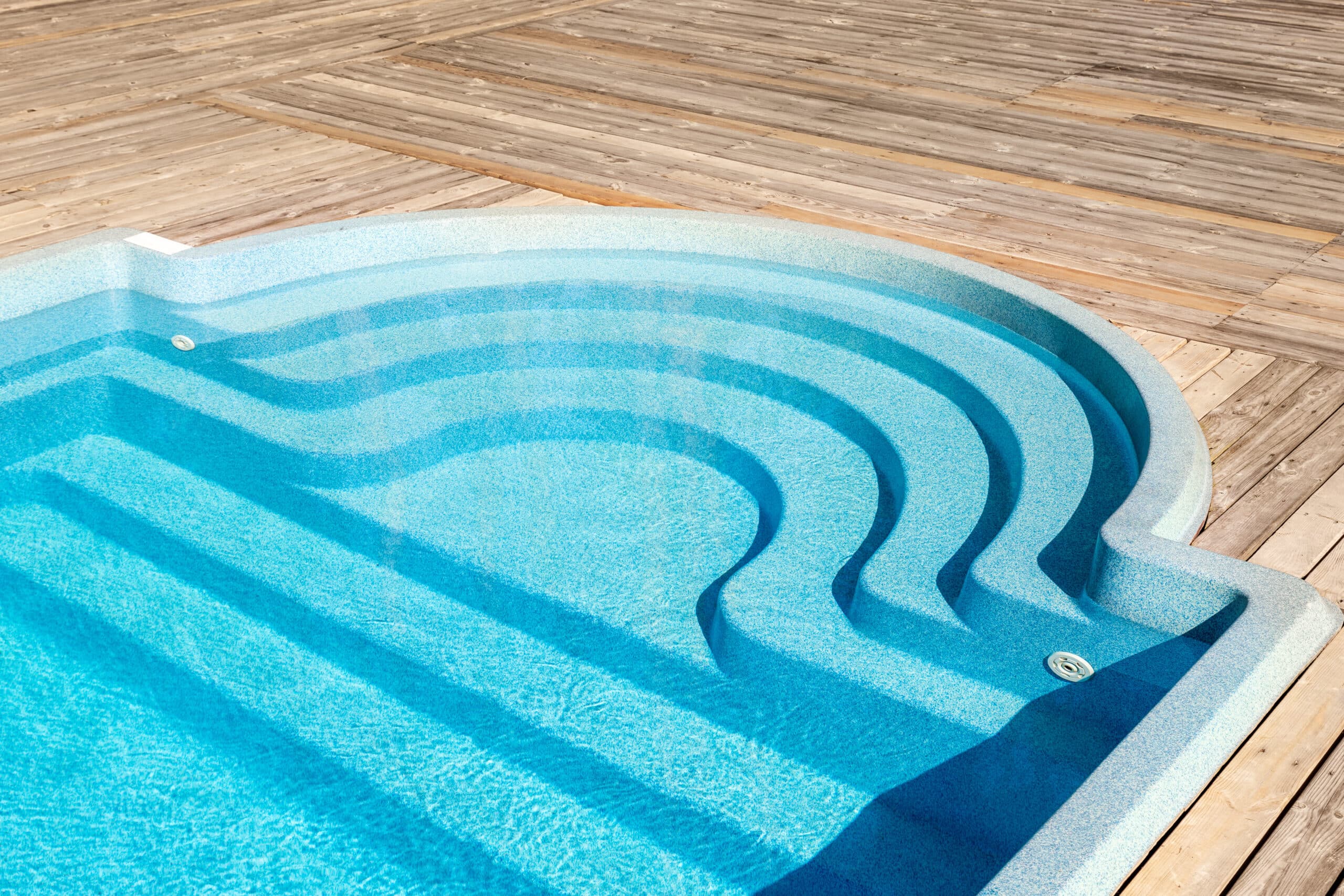 New modern fiberglass plastic swimming pool entrance step with clean fresh refreshing blue water on bright hot summer day at yard or resort hotel spa area. Wooden flooring deck of teak or larch board Swimming Pools 6 | AdobeStock 410082526 1 scaled